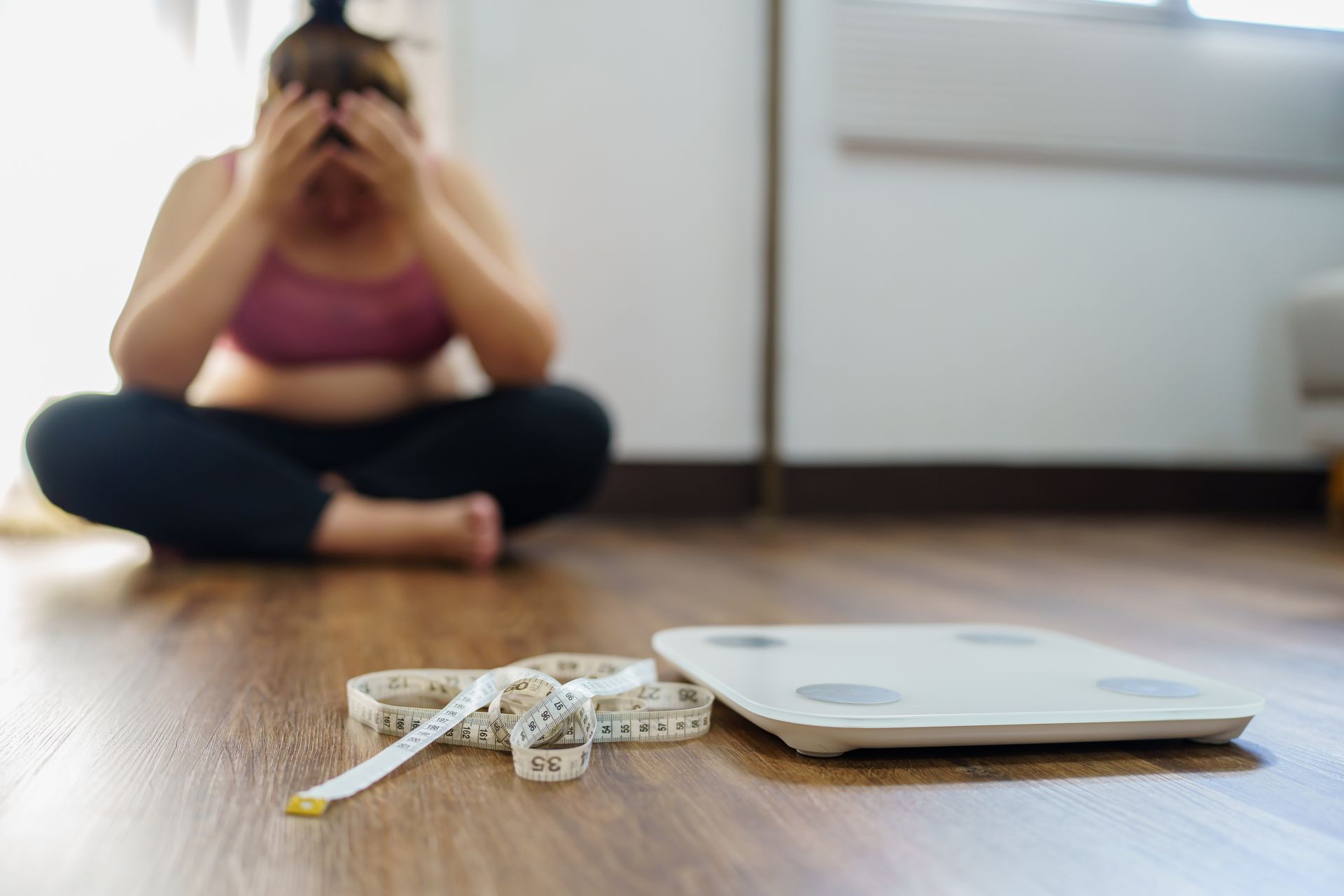 Person sitting cross-legged on a floor beside a scale and measuring tape, looking stressed