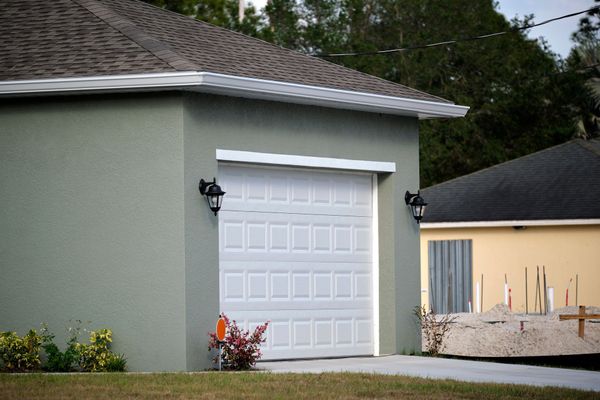 White garage door on a light green house beside a beige building and driveway, with shrubs and trees nearby