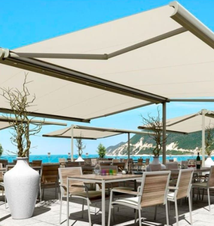 Restaurante al aire libre con toldos retráctiles beige, mesas, sillas y vista a la costa.