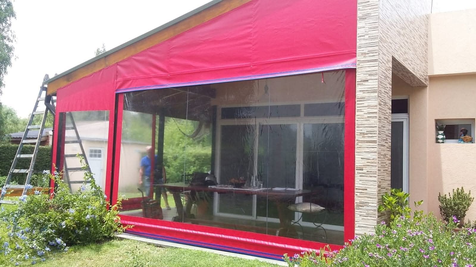 Toldo rojo con laterales de vinilo transparente que encierra un patio con puertas blancas.