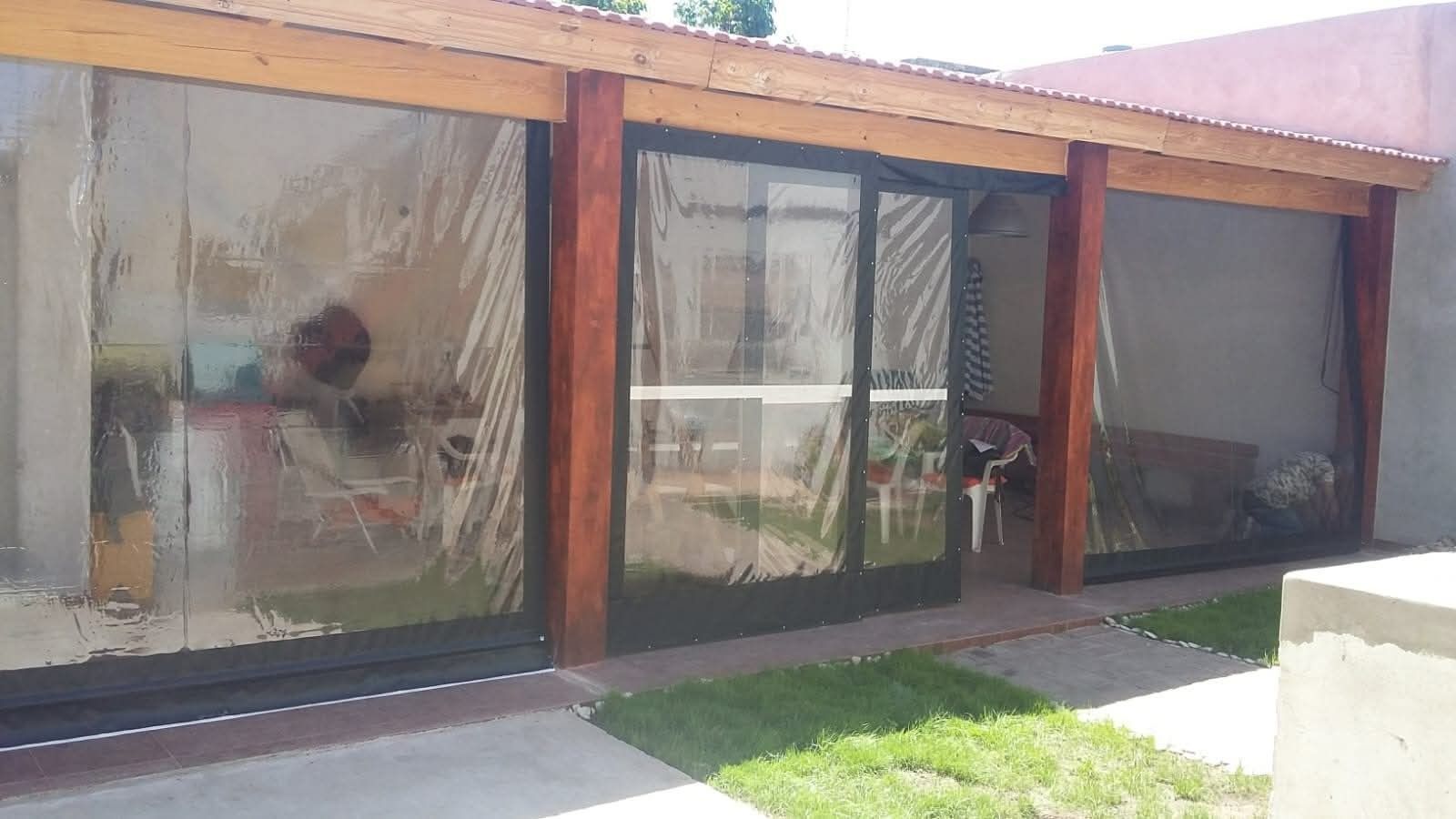 Patio cubierto con cortinas de plástico transparente, postes de madera y puerta corrediza de vidrio.