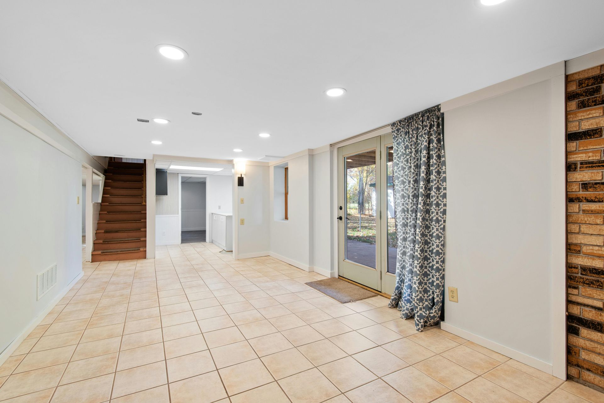 Empty basement room with light-colored tile floor, door to the outside, exposed brick wall, and a staircase.