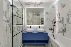Modern bathroom with blue vanity, two white sinks, marble walls, and glass shower.