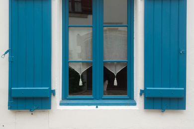Blue shutters flanking a blue-framed window with sheer curtains; set in a white wall.