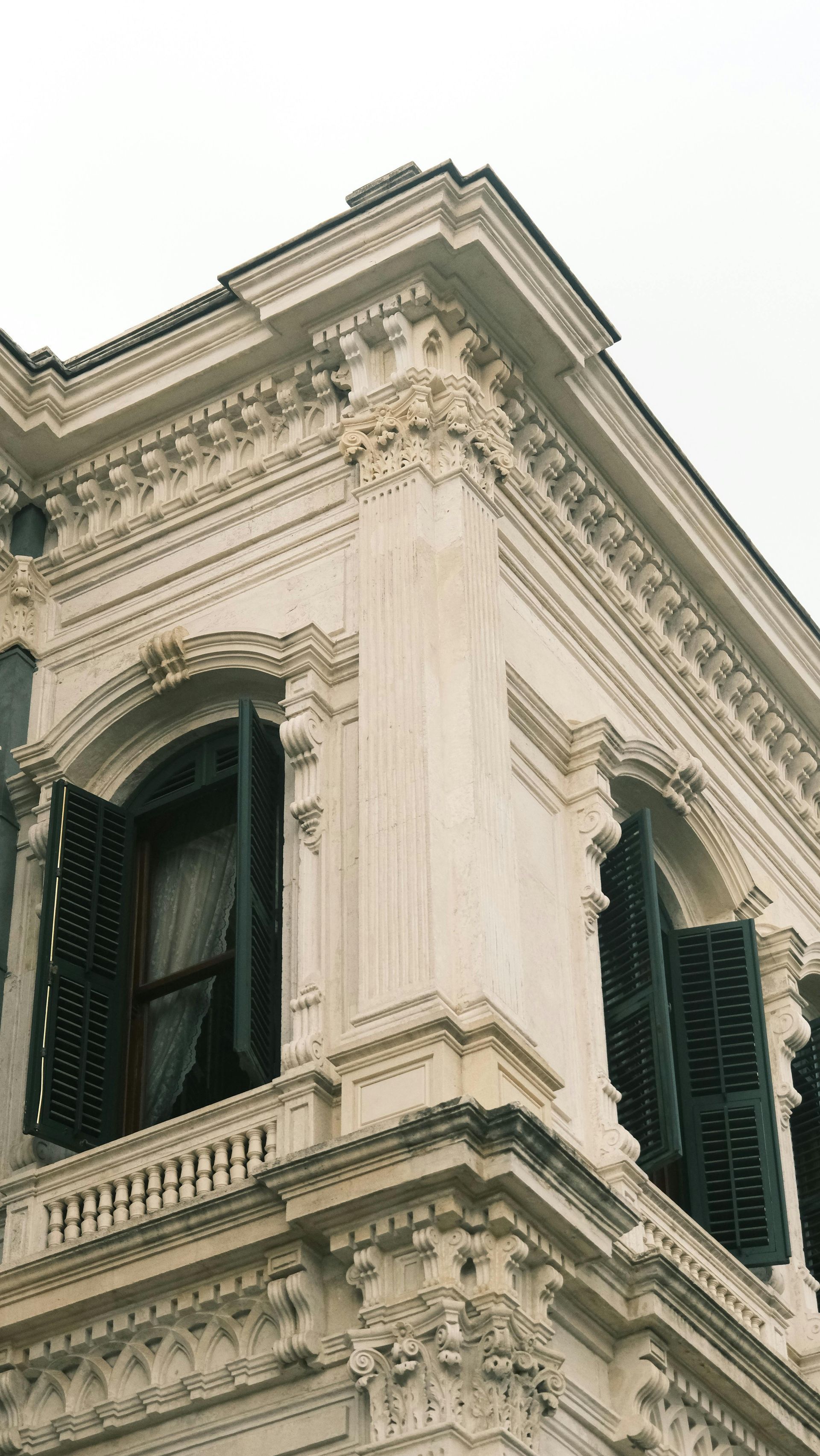 Corner of a light-colored building with ornate details, arched windows, and dark green shutters.