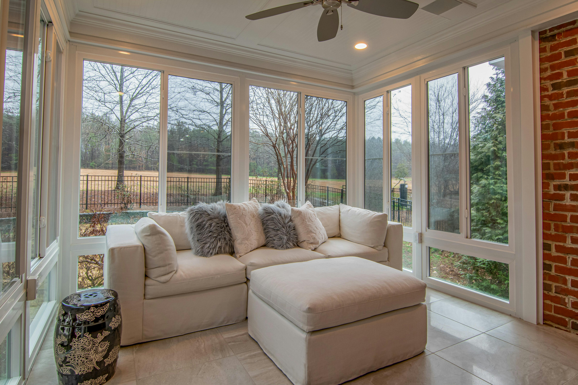 Sunroom with white sectional sofa and ottoman, surrounded by windows with outdoor view and brick wall.