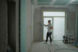 Man spraying a wall in a room under renovation with drywall mud. The walls are unfinished, with a ladder and equipment.