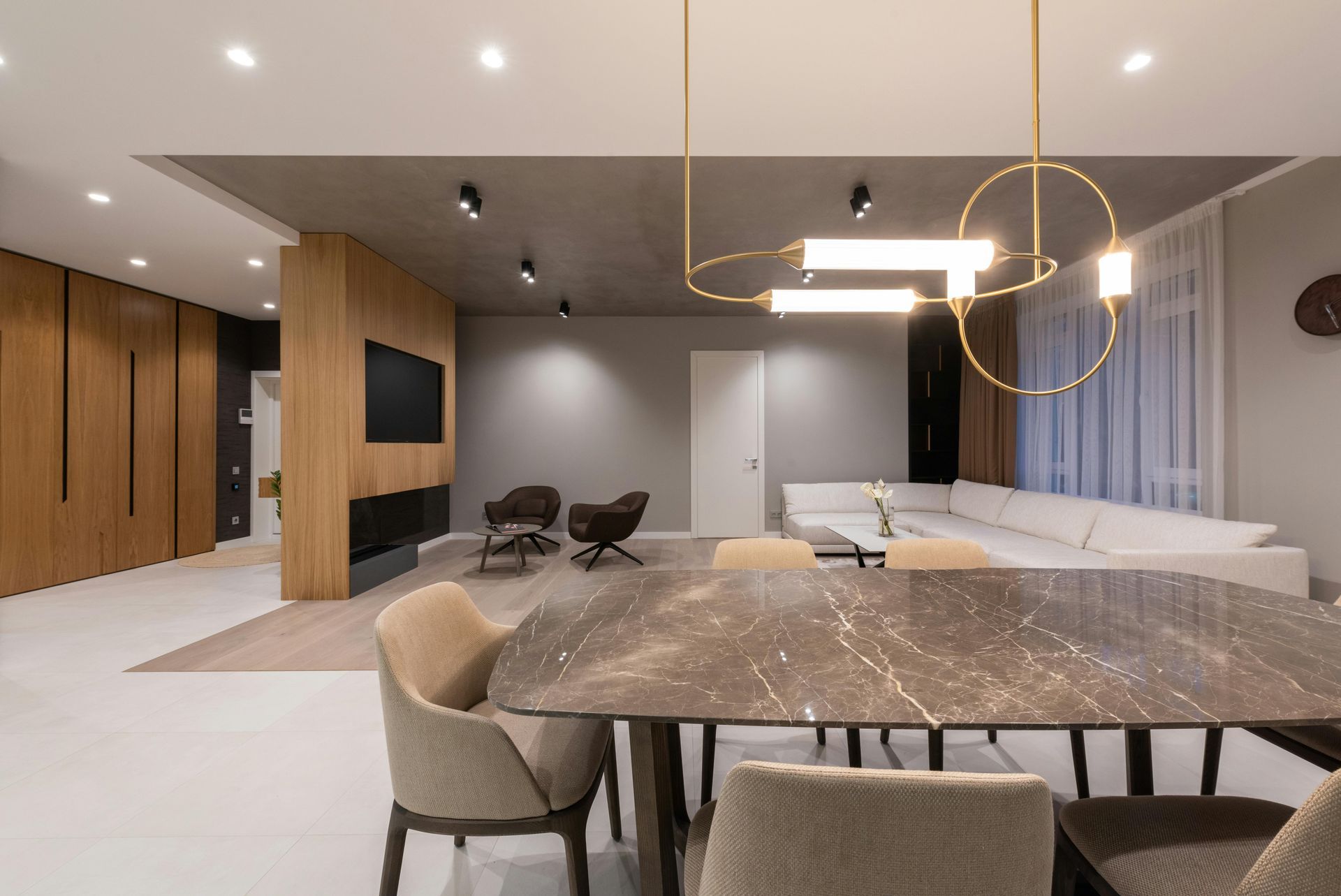 Modern dining room with marble table, chairs, and gold chandelier. Living area with sofa, TV, and wood paneling.