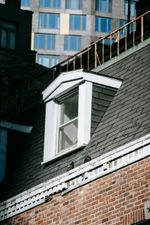 Brick building with a dormer window, black shingle roof, and a modern building in the background.