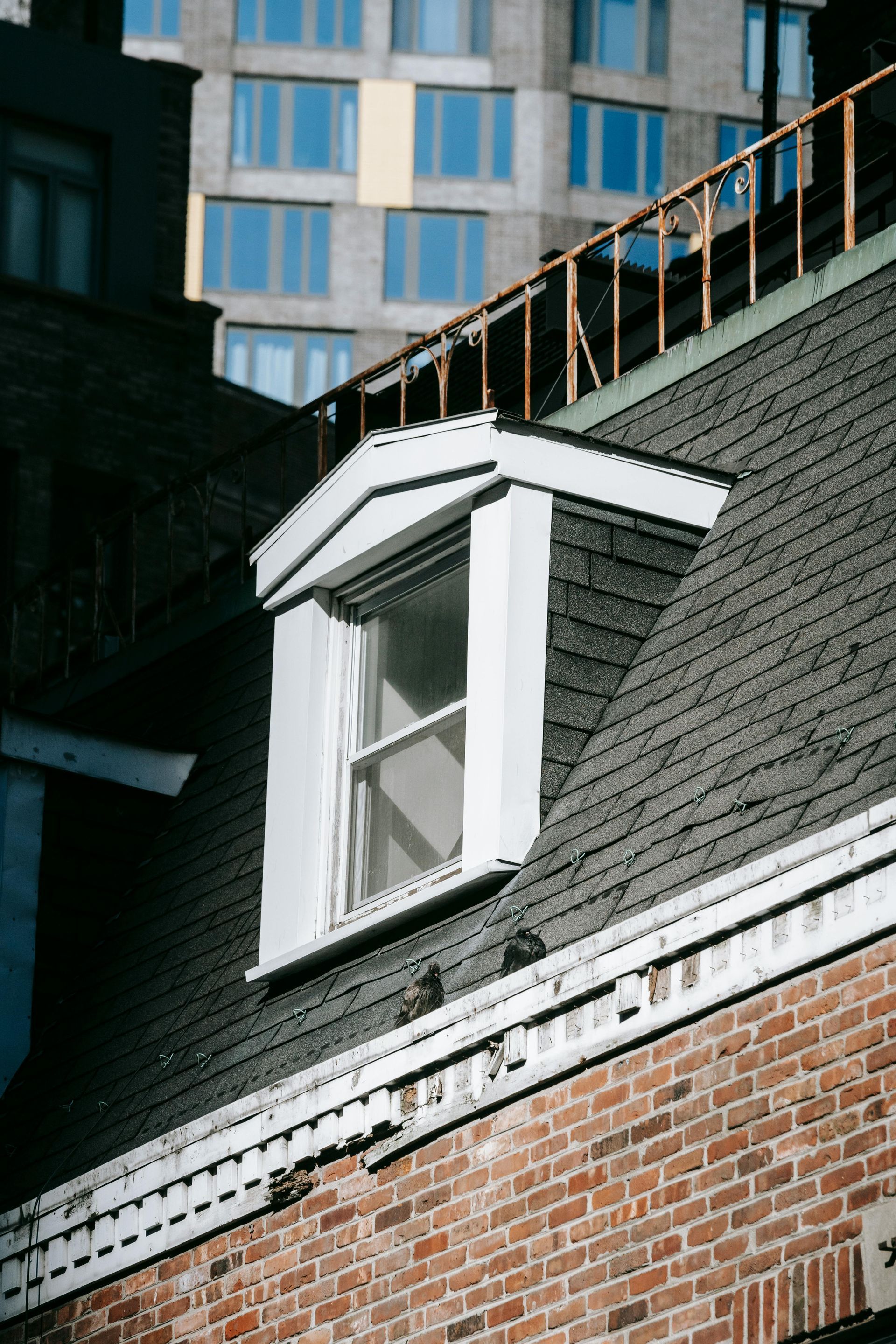 Brick building with a dormer window, black shingle roof, and a modern building in the background.