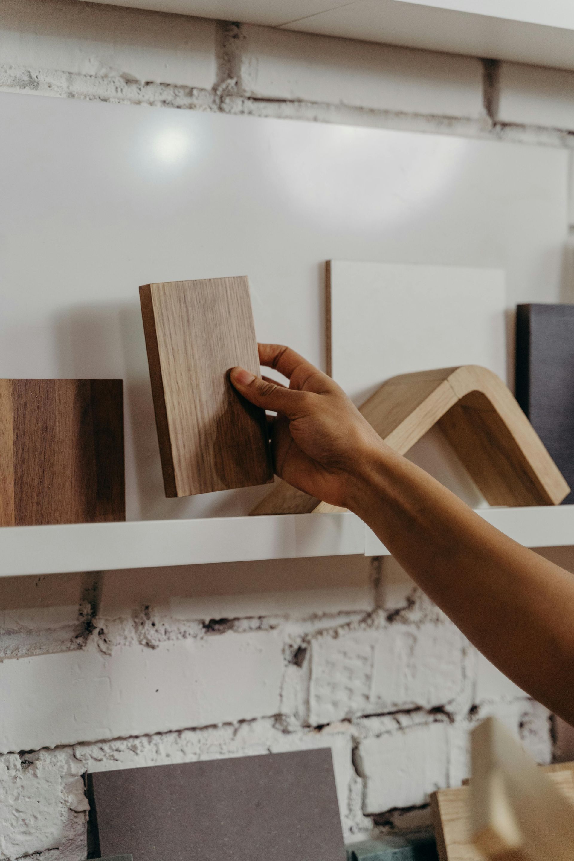 Hand holding a wooden sample from a shelf displaying various material swatches. White brick wall backdrop.
