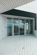 White building with glass entrance, black awning, and light-colored wooden deck. Overcast sky visible through the windows.