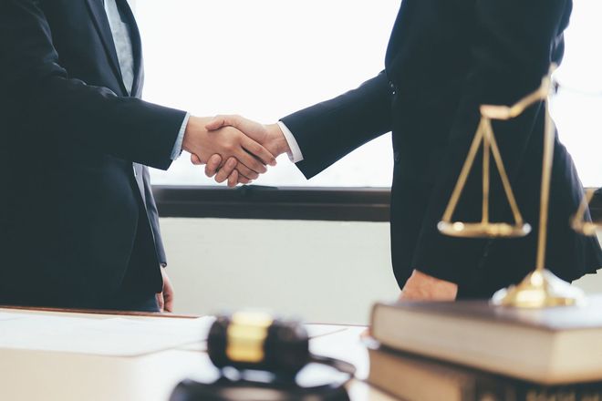 Two businesspeople shaking hands across a desk with a justice scale in the foreground. Two businesspeople shaking hands across a desk with a justice scale in the foreground.