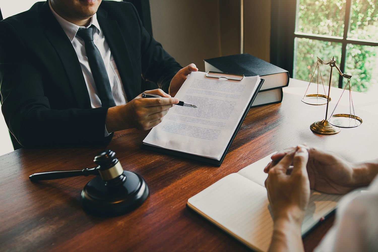 Lawyer discussing a case at a desk with documents, gavel, and scales by a window Lawyer discussing a case at a desk with documents, gavel, and scales by a window