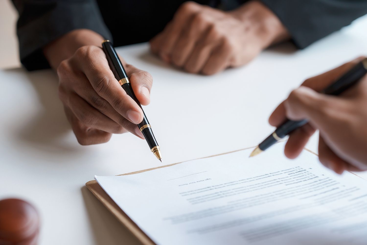 Two hands signing a document with pens over a clipboard on a desk Two hands signing a document with pens over a clipboard on a desk