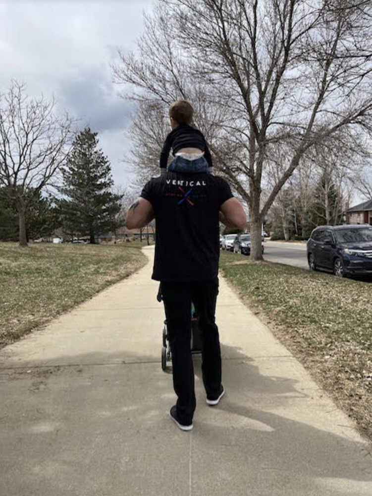 A man with a child on his shoulders walks along a path, pushing a stroller. Cloudy day, park setting.