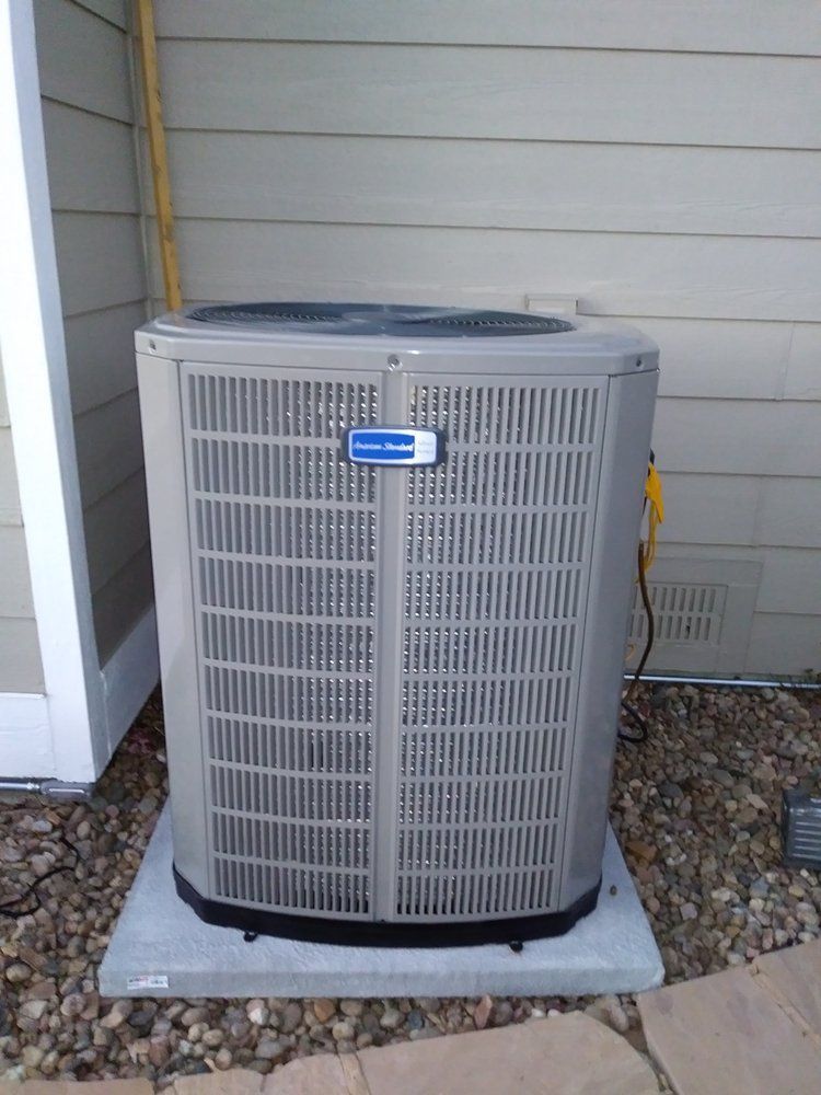 An air conditioning unit on a concrete pad against a tan siding wall, brown gravel ground.
