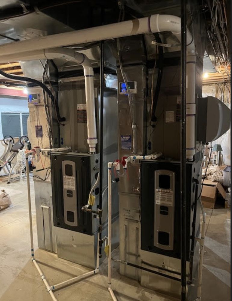Two furnaces side-by-side in a basement, with white PVC pipes and black wiring.