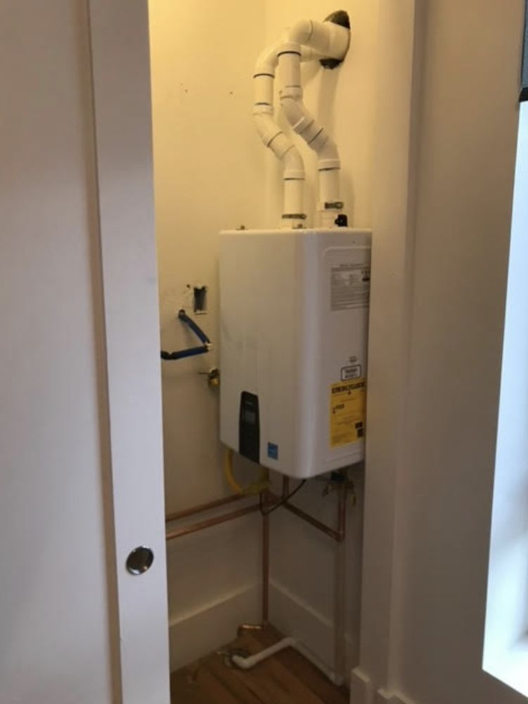 White tankless water heater in a small closet with white vent pipes.
