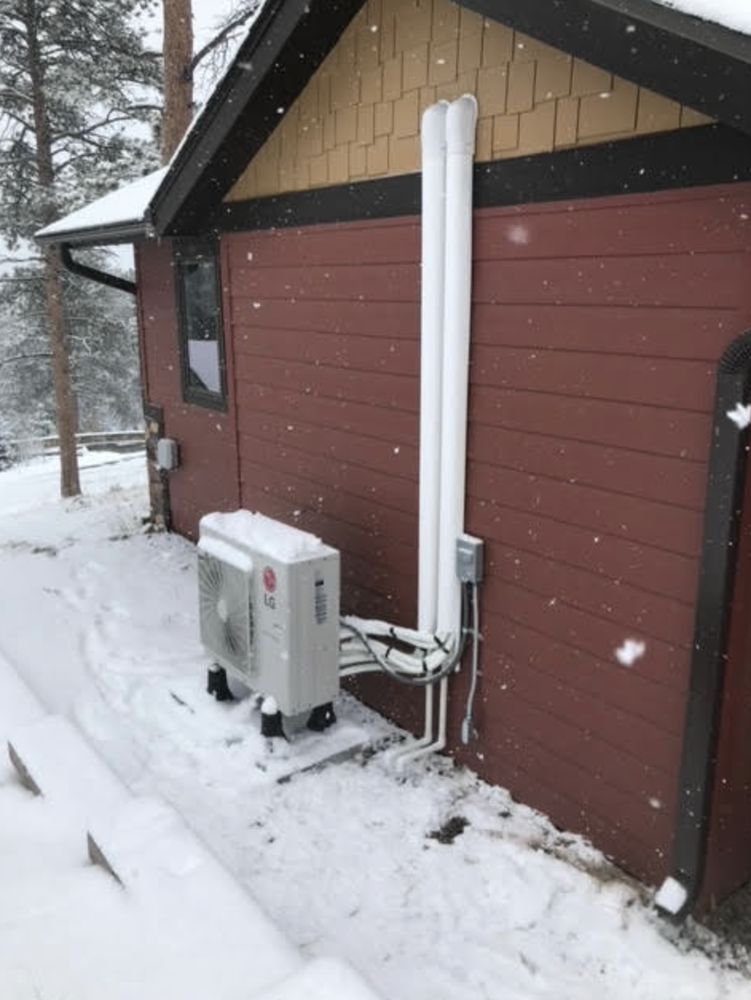 Outdoor heat pump unit next to a building, covered in snow, with insulated pipes running vertically.