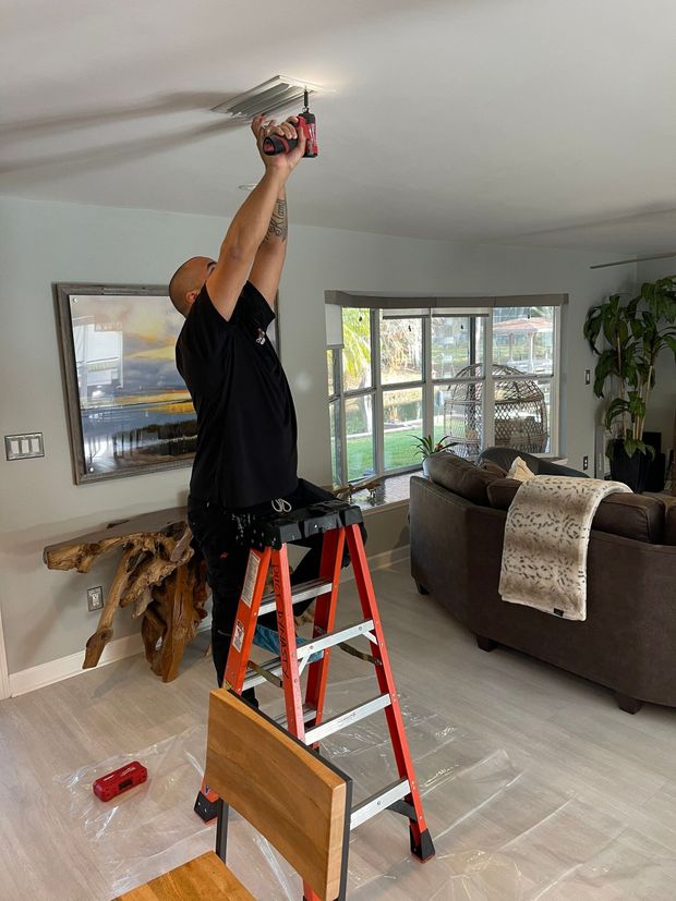 A man is standing on a ladder in a living room holding a drill.