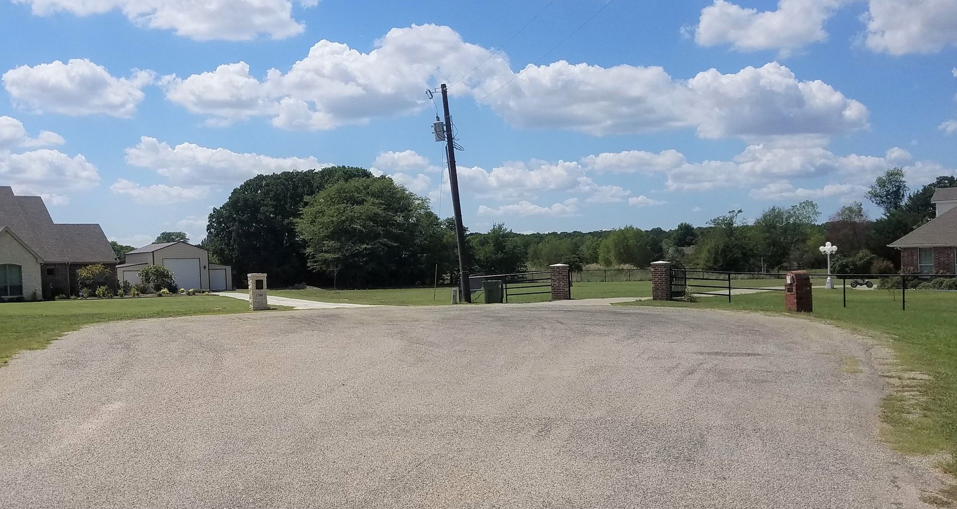 Gravel driveway leads towards houses and trees under a blue sky with fluffy clouds.