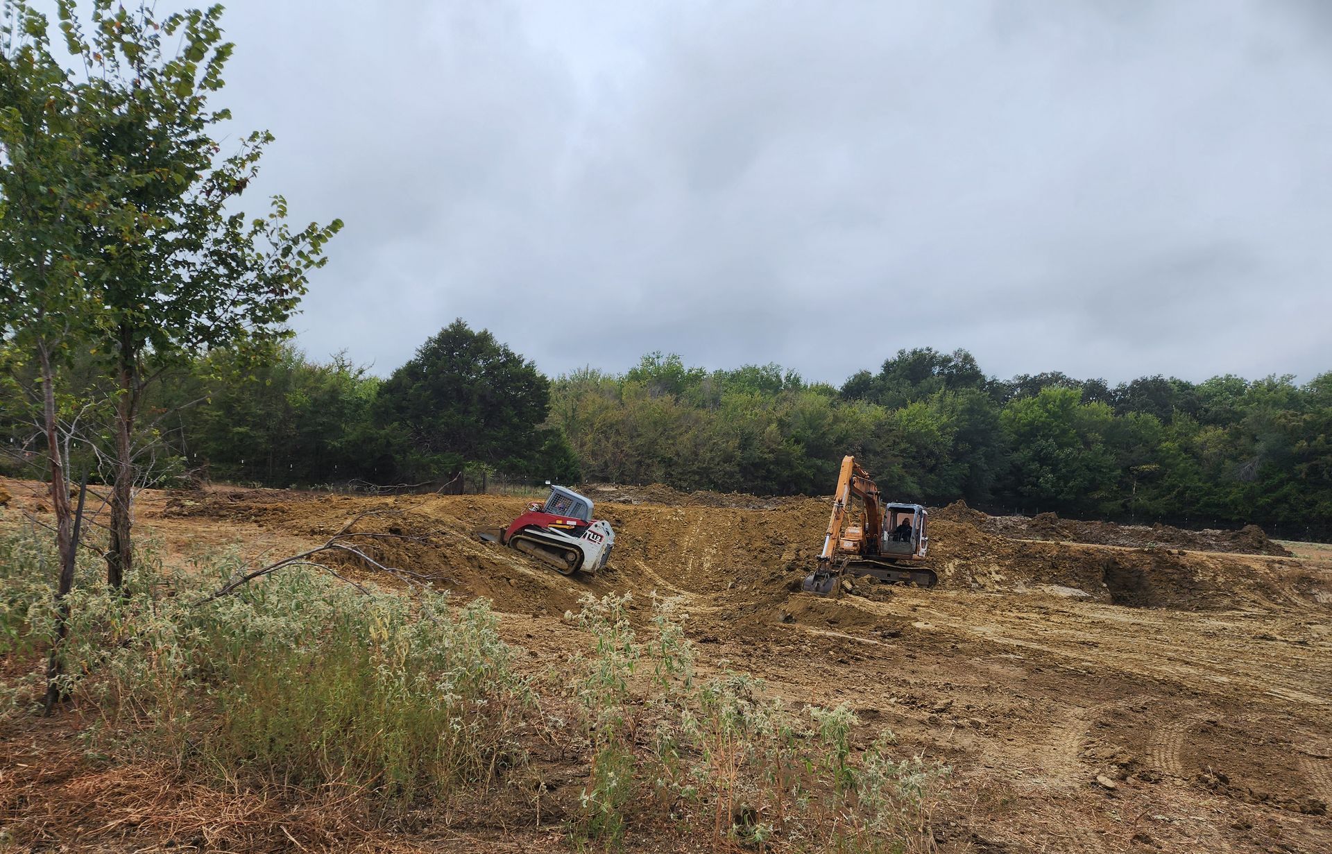 Two excavators work on a dirt clearing under a cloudy sky, surrounded by trees and scrub.