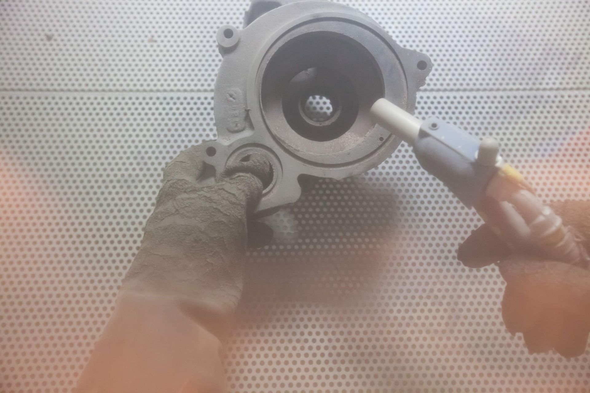 A person wearing a protective glove sandblasts the interior of a circular metal engine component.