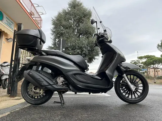 Uno scooter nero parcheggiato su una strada asfaltata con bauletto, parabrezza e cavalletto montati.