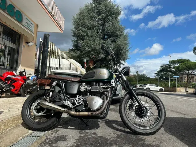 Una motocicletta Triumph verde scuro parcheggiata all'angolo di una strada, con una motocicletta rossa visibile sullo sfondo.