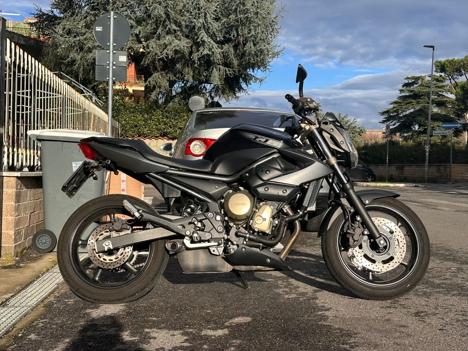 Una motocicletta Yamaha XJ6 nera parcheggiata su un piazzale asfaltato accanto a un edificio e a un cestino dei rifiuti in una giornata di sole.