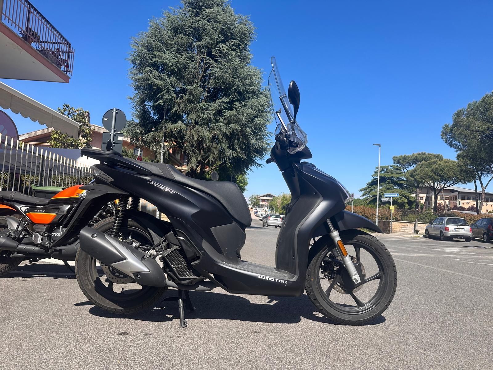Scooter nero parcheggiato in una strada soleggiata con alberi ed edifici sullo sfondo.