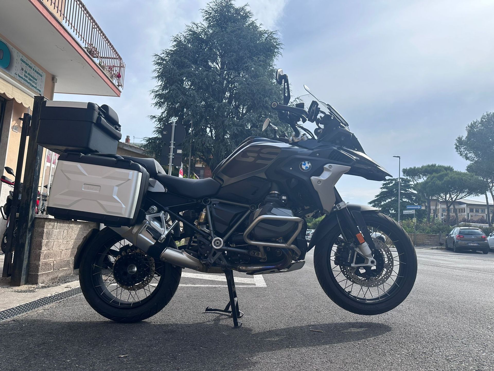 Una motocicletta da avventura nera parcheggiata su una strada accanto a un edificio, con alberi e cielo nuvoloso sullo sfondo.