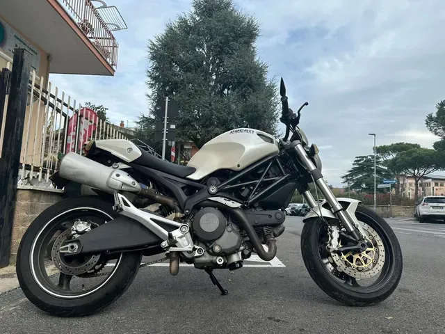 Una motocicletta Ducati Monster bianca parcheggiata su una superficie asfaltata, con un albero e un edificio sullo sfondo.