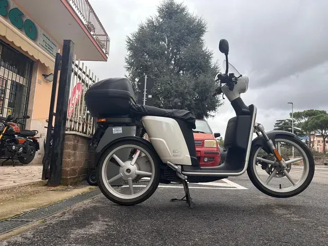 Uno scooter bianco e nero parcheggiato su una strada asfaltata con un box portaoggetti nero sul retro.