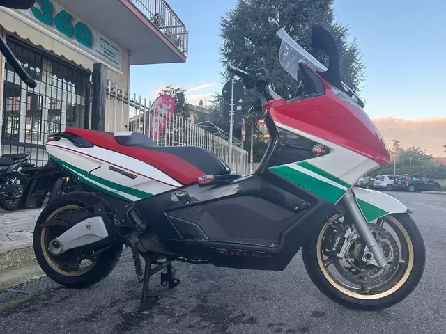Vista laterale di uno scooter Gilera GP800 rosso, bianco e verde parcheggiato su un piazzale asfaltato vicino a un edificio.