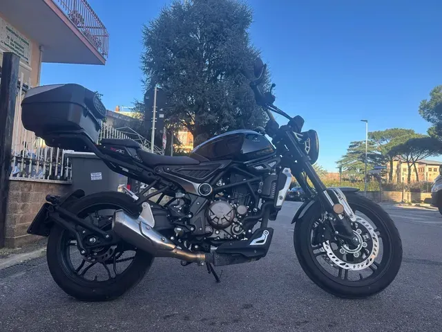 Una motocicletta nera con bauletto posteriore parcheggiata su una strada asfaltata sotto un cielo azzurro e limpido.