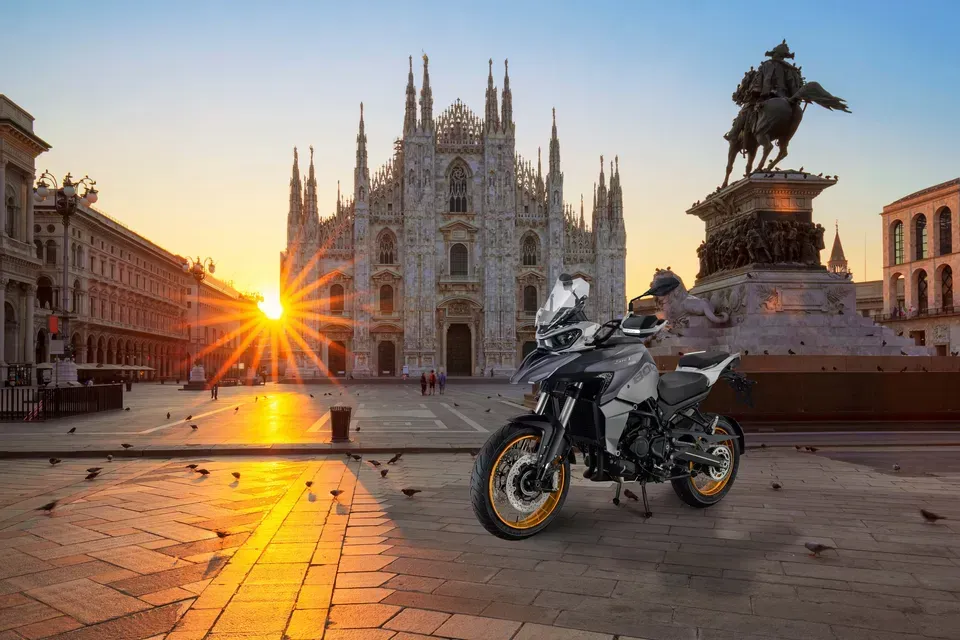 Una motocicletta parcheggiata in una piazza all'alba, con il Duomo di Milano e una statua sullo sfondo.