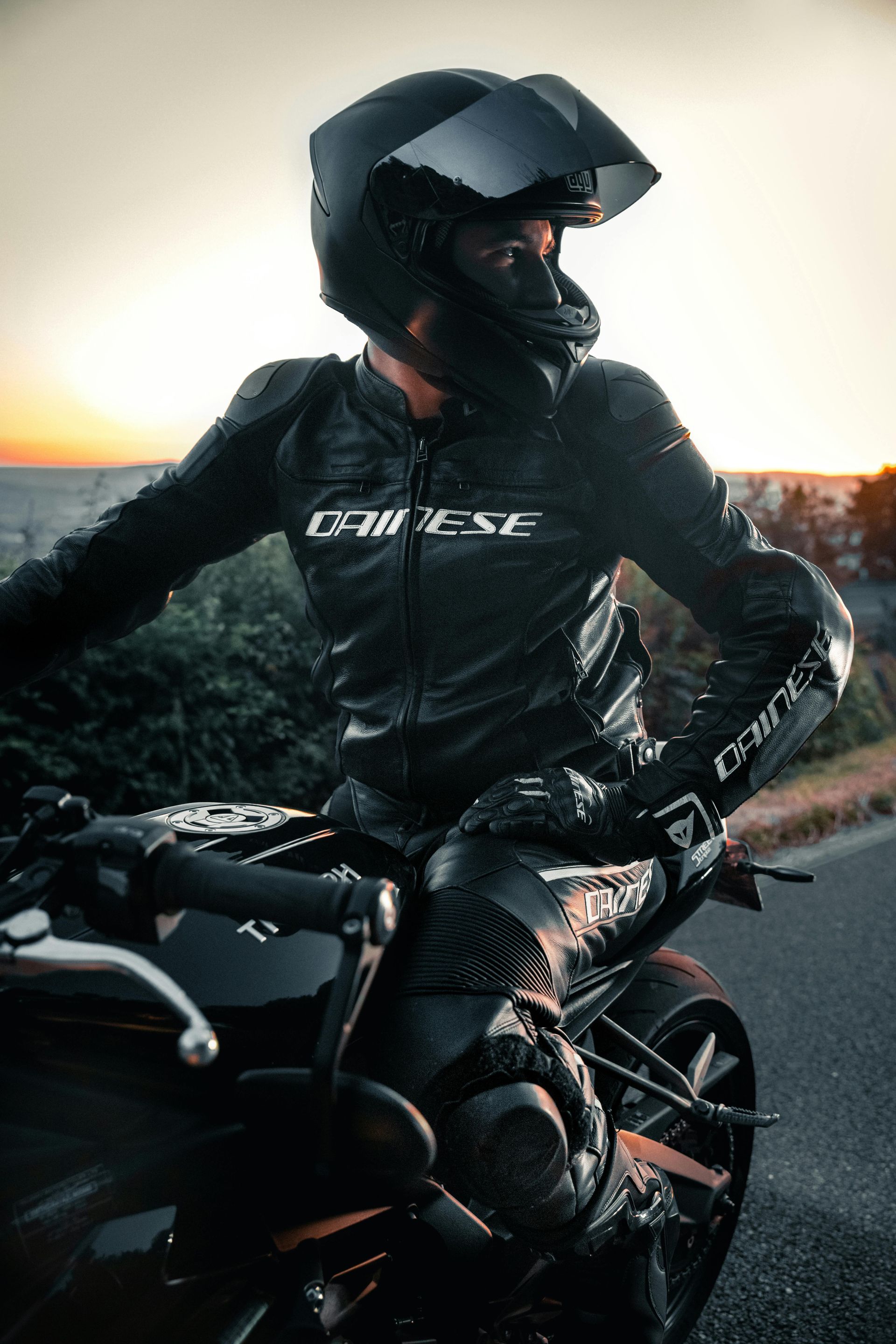 Un motociclista con abbigliamento Dainese nero e casco è seduto sulla sua moto su una strada al tramonto.