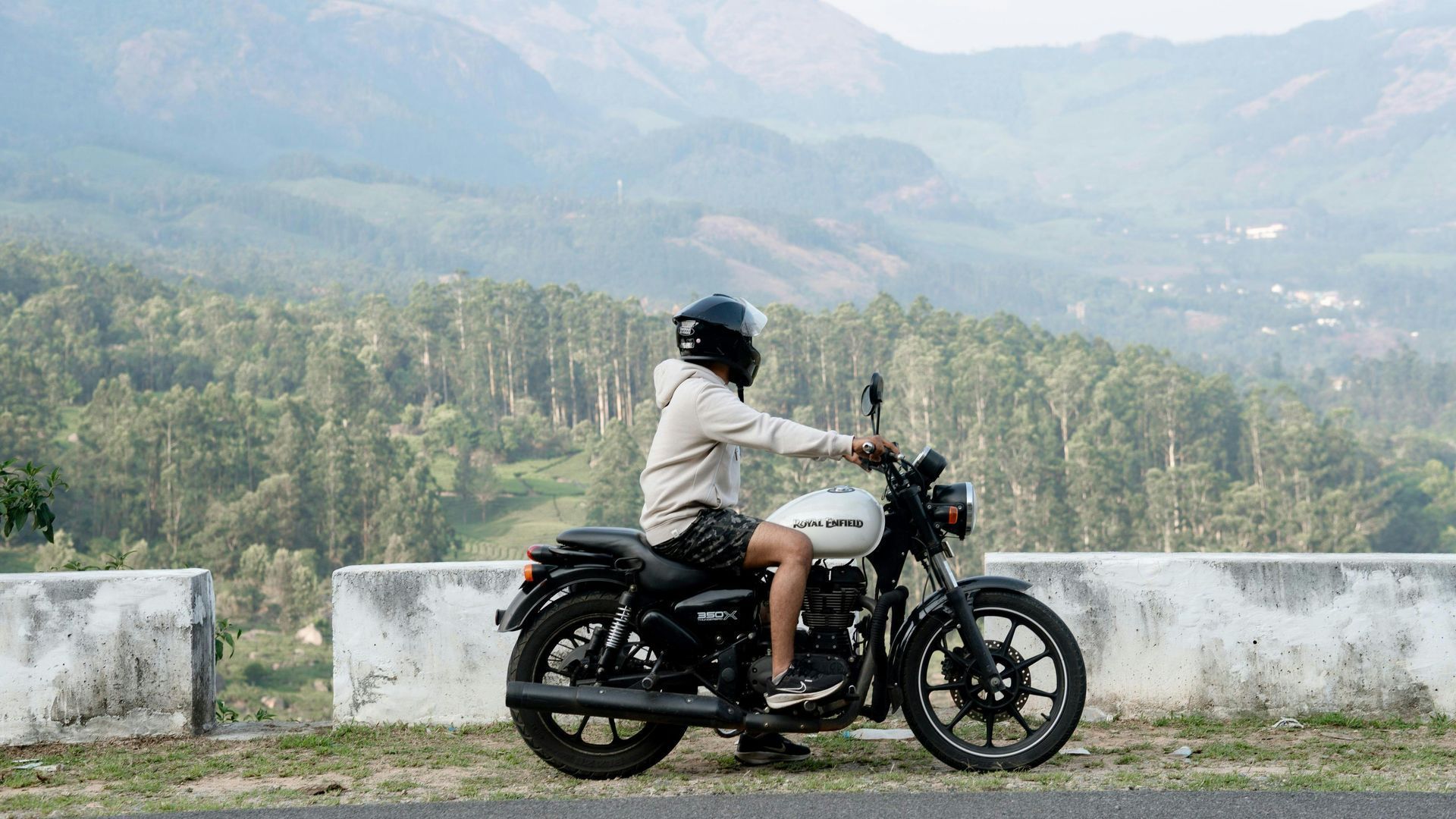 Una persona con casco e felpa leggera è seduta su una motocicletta bianca parcheggiata vicino a una barriera di