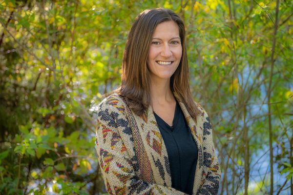 Woman with brown hair smiles, wearing patterned shawl over black shirt, outdoors with blurred greenery.