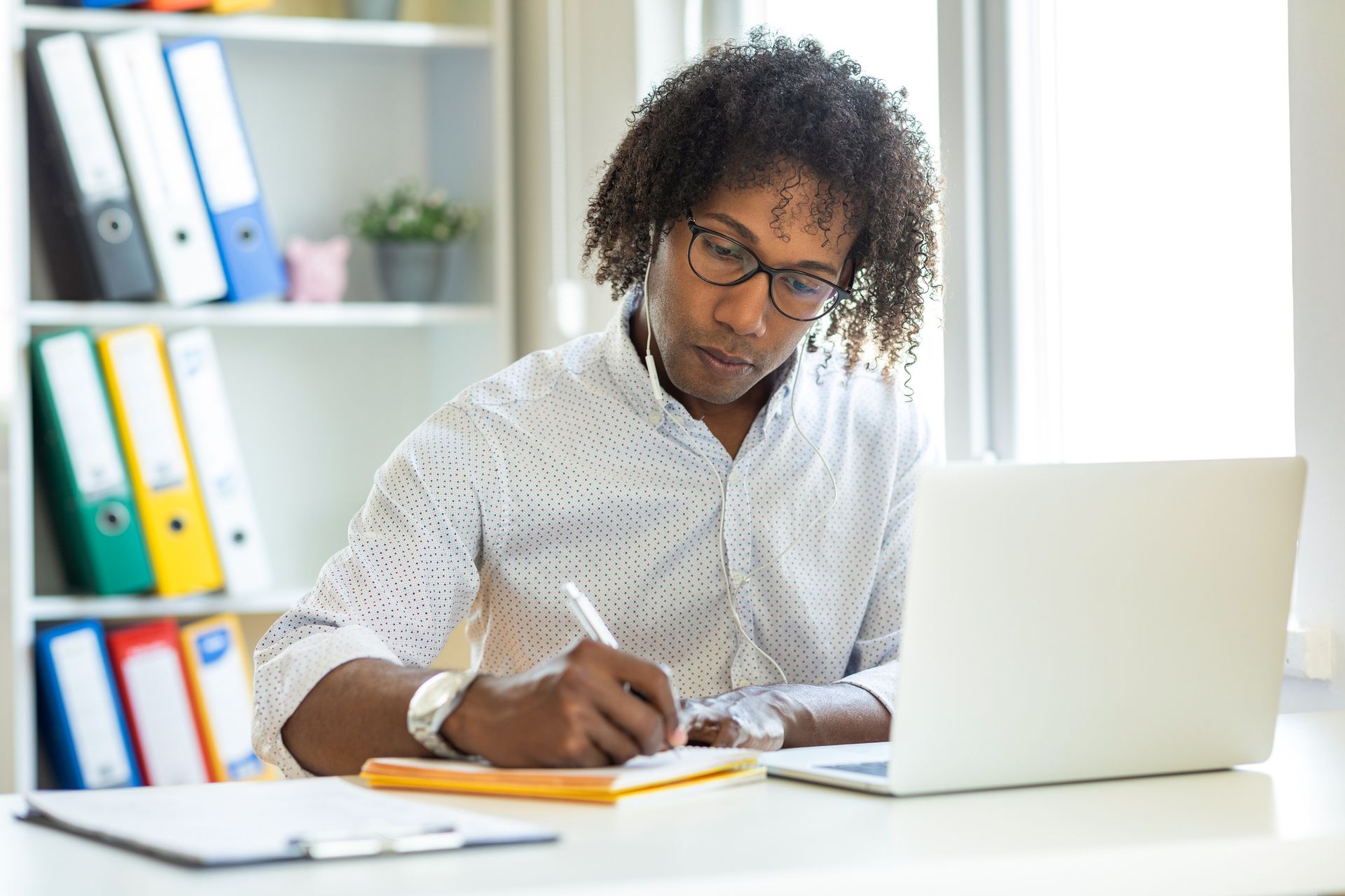 A man is sitting at a desk using a laptop computer and writing in a notebook.