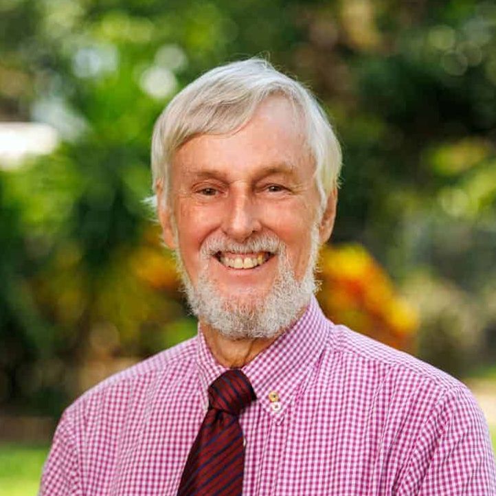 A smiling older man with white hair and a beard, wearing a pink checkered shirt and a burgundy tie, posing outdoors.