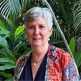 Woman with short gray hair, wearing a colorful patterned shawl, smiles outdoors in front of leafy green plants.