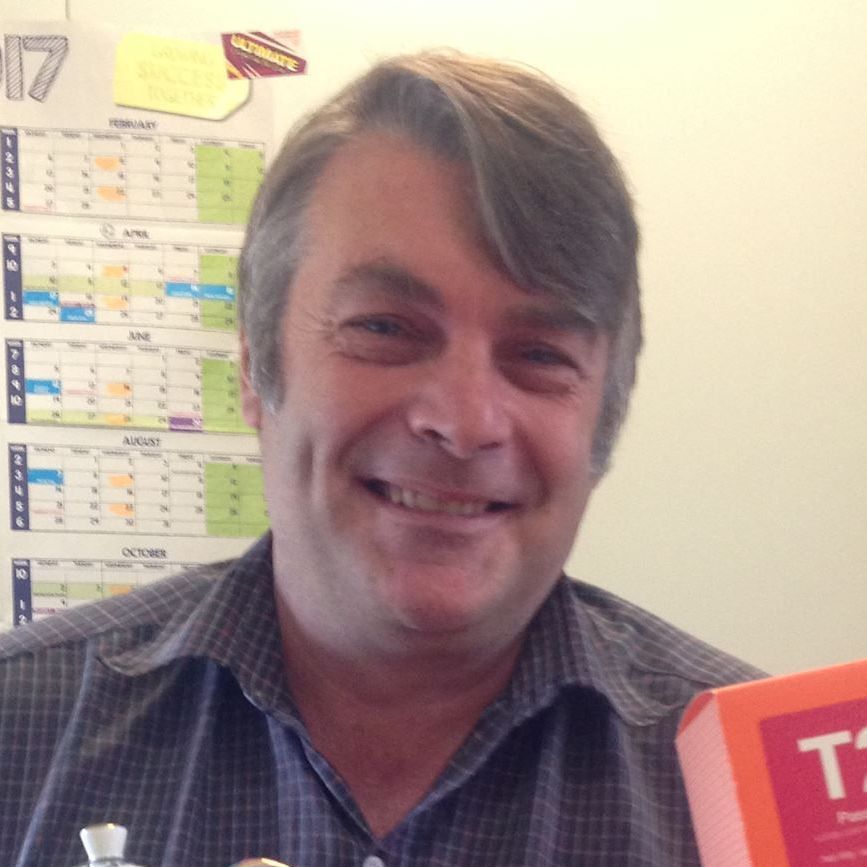 A smiling man with gray hair, holding a small orange box, in front of a calendar.