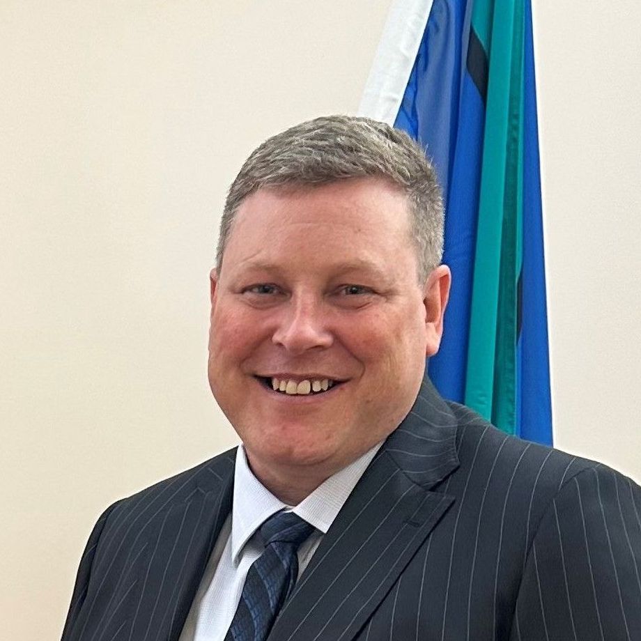 A smiling man in a suit and tie stands in front of a flag with blue, green, and white stripes.