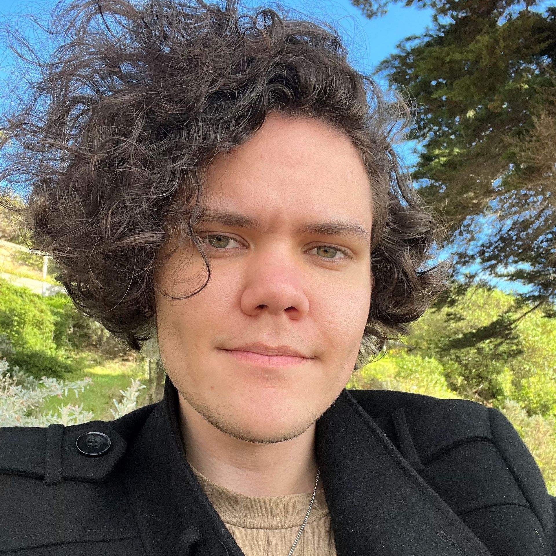 Young man with curly brown hair in front of greenery and trees. He smiles, wearing a dark jacket and tan shirt.