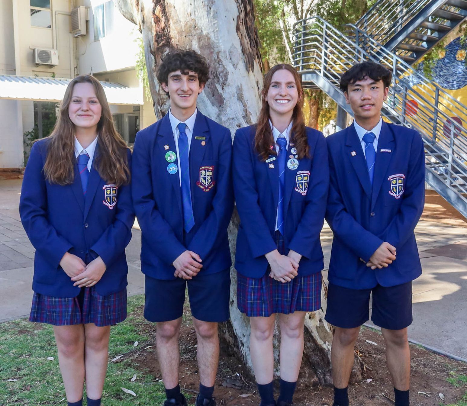 Four students in school uniforms pose in front of a tree, smiling. They wear navy blazers, ties, and either plaid skirts or navy shorts.