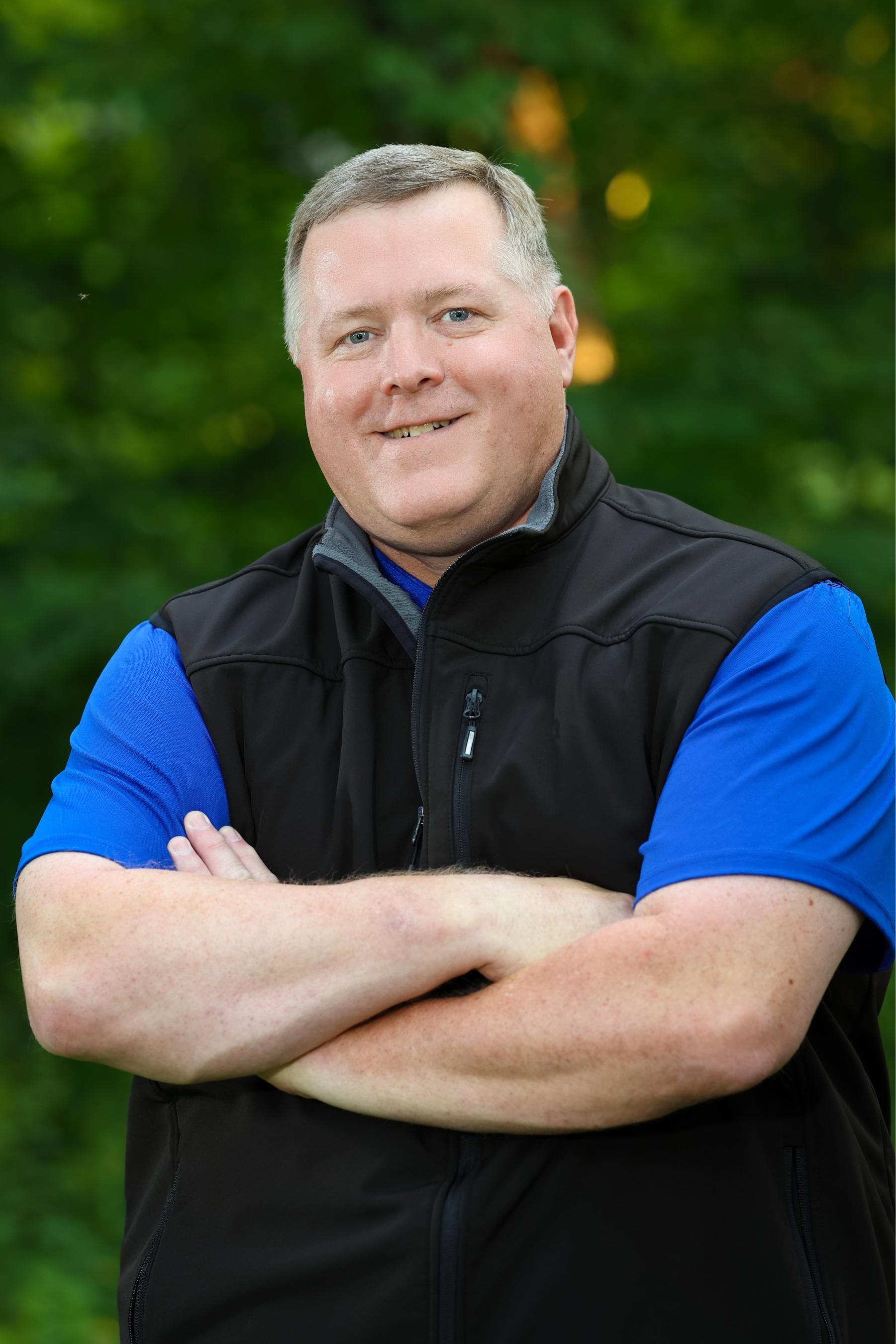 A man wearing a blue shirt and a black vest is standing with his arms crossed.