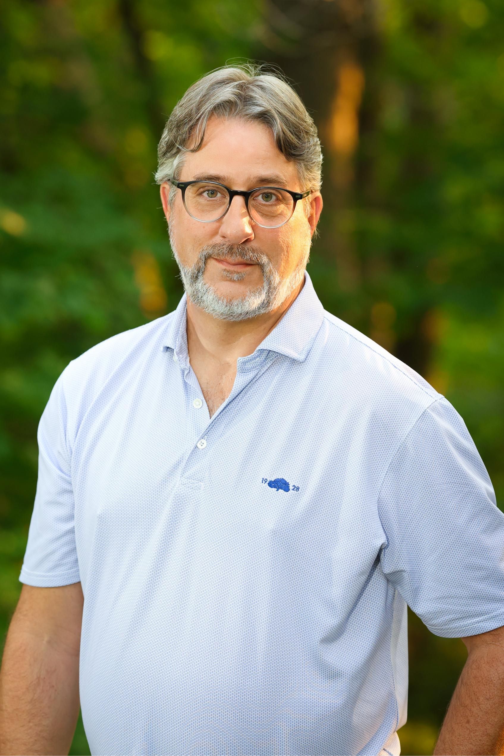 A man with a beard and glasses is standing in front of a forest.
